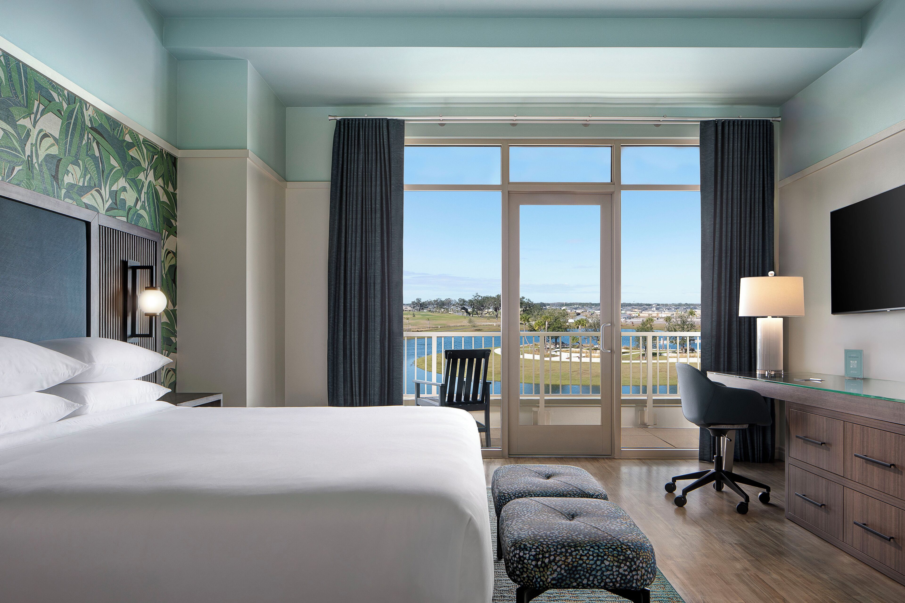 Habitación de hotel con una cama grande, un escritorio, una silla, televisión y un balcón con vistas.