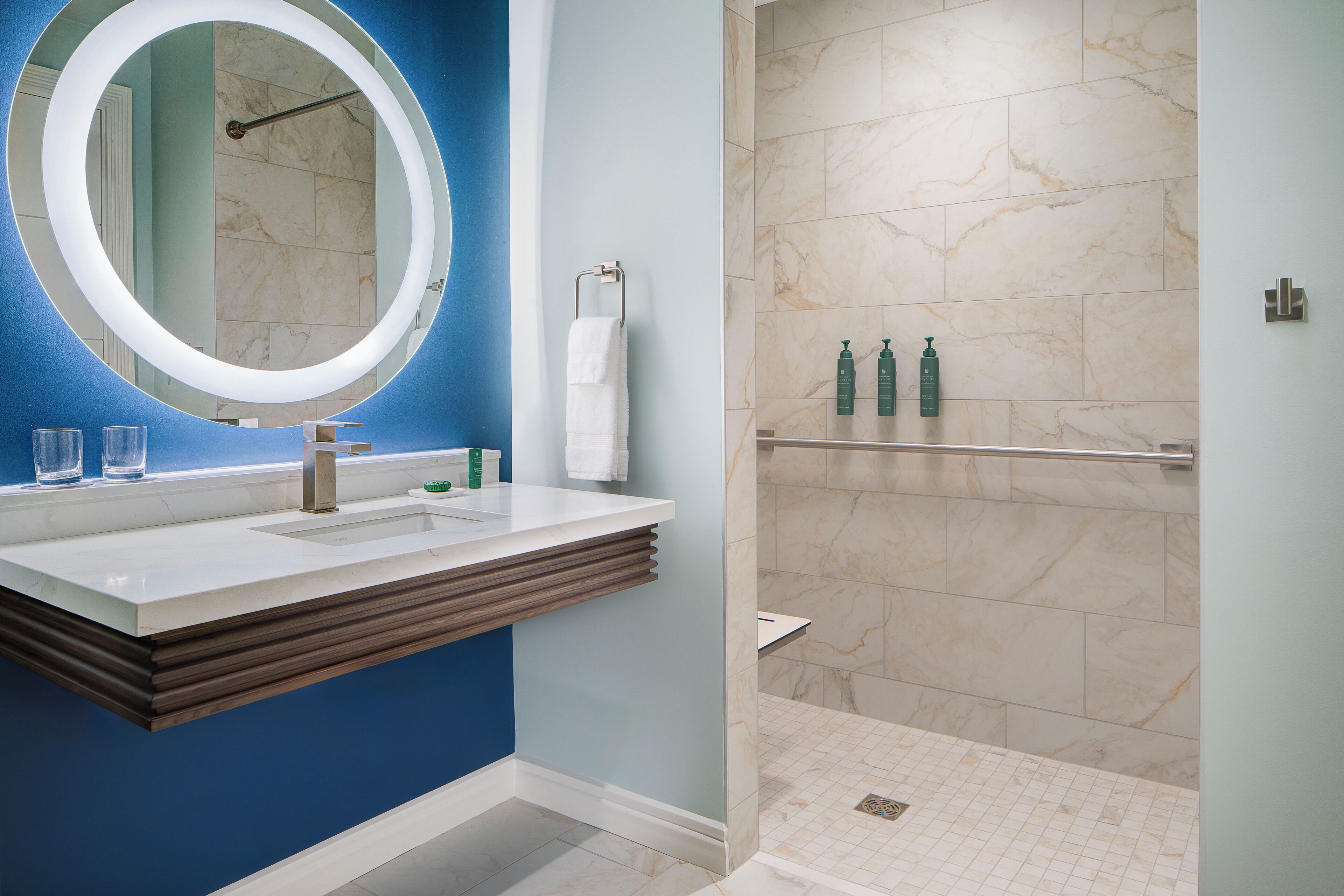 Une salle de bain moderne avec un mur bleu, un miroir rond, un meuble-lavabo en bois et un espace douche aux carreaux de marbre.