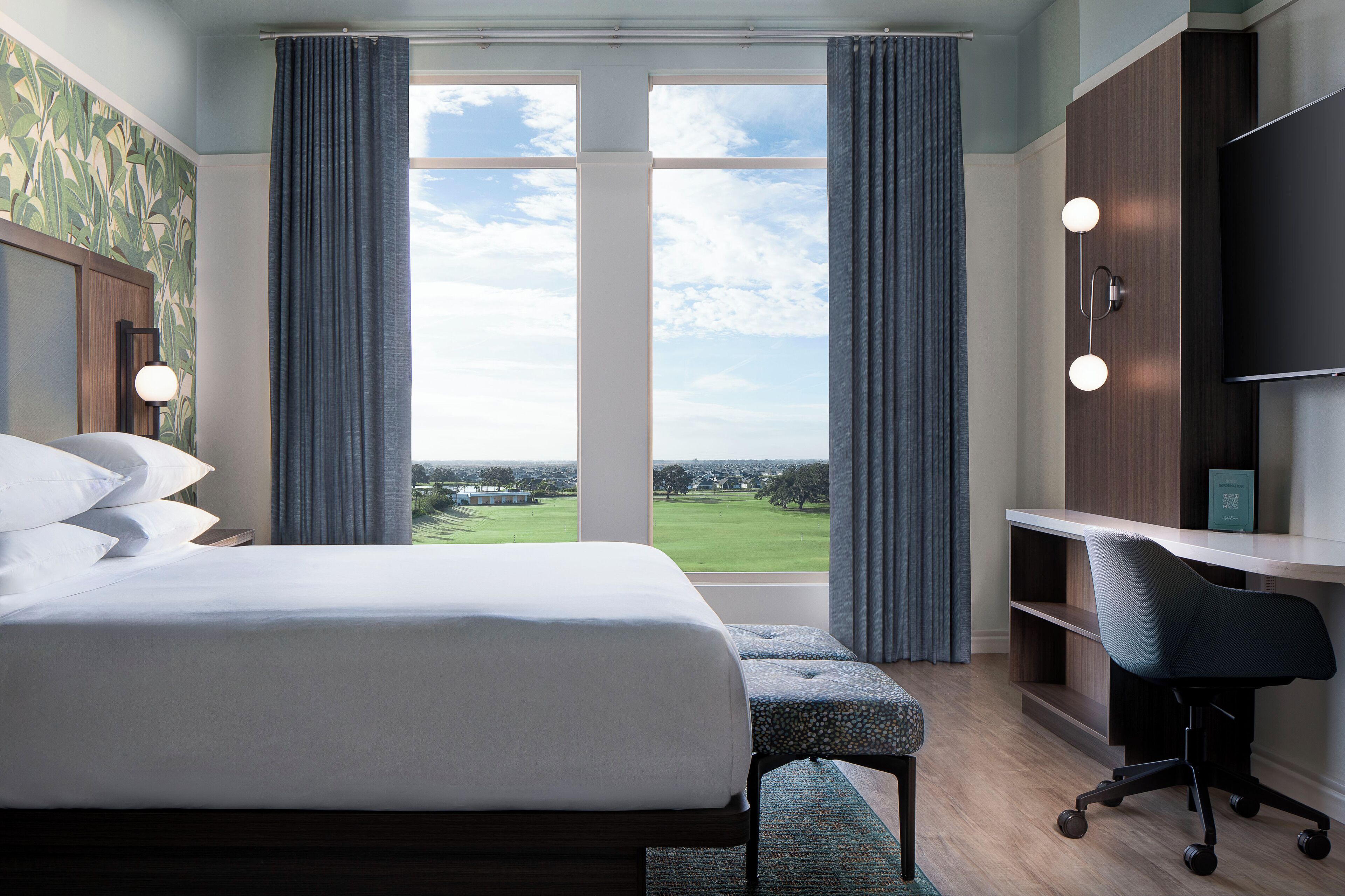Habitació d'hotel amb un llit gran, un escriptori amb una cadira i vista a un camp de golf per la finestra.