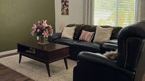 Uma sala de estar com parede verde escura, poltrona de couro preto, sofá preto, mesa de centro de madeira e luminária de chão.