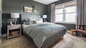 Un dormitorio con una cama, mesitas de noche, una lámpara, un ciervo montado en la pared y una ventana con cortinas.