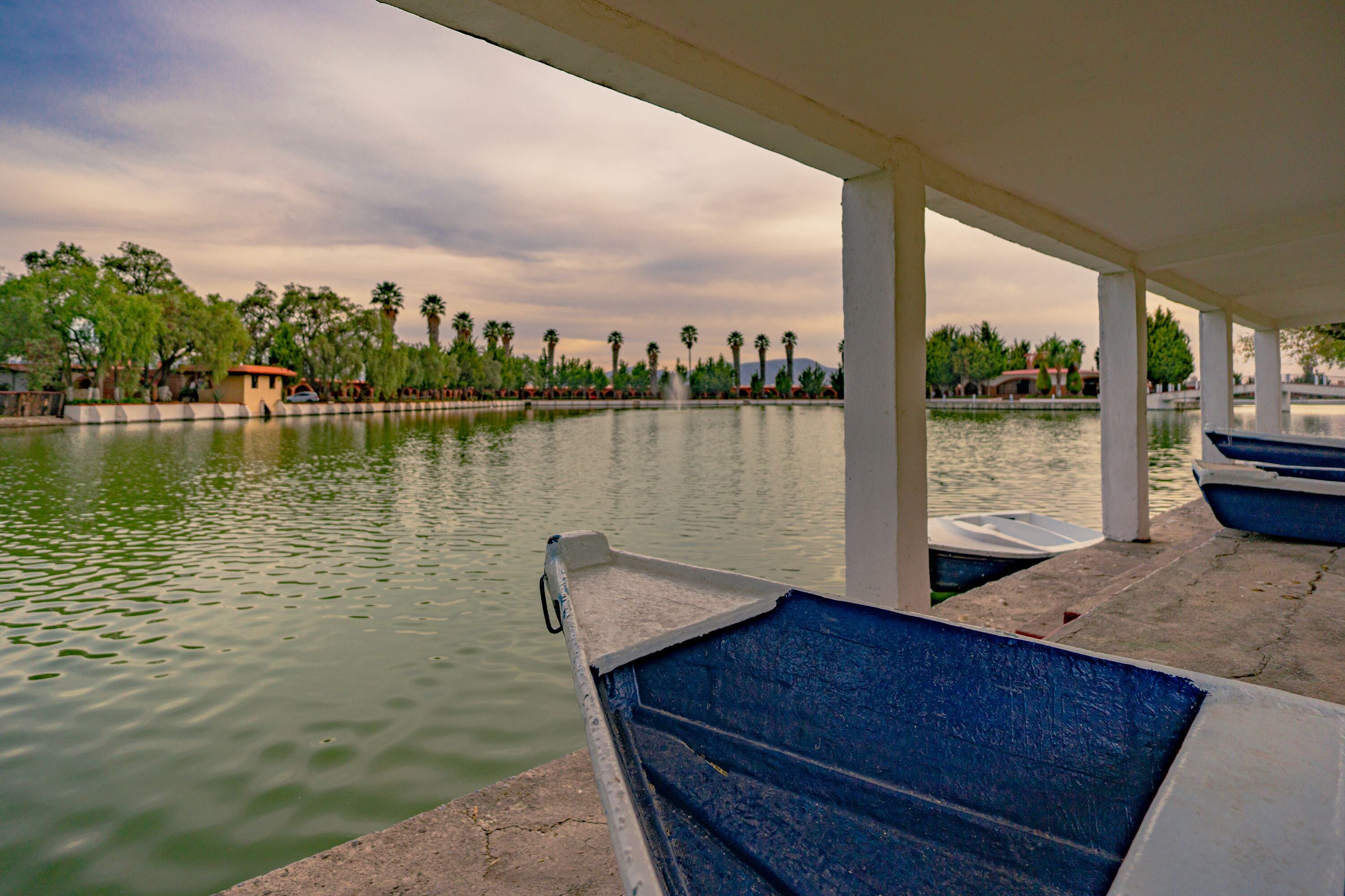 A calm water body with boats docked, a covered area with columns, and a row of trees and buildings in the background.