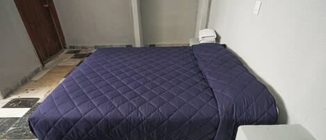 A bed with a quilted cover, a wooden door, a tiled floor, and a white wall with a power outlet.