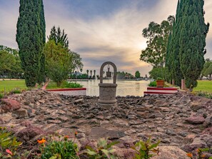 Una fuente de piedra con un arco central, rodeada de terreno rocoso y vegetación, con un cuerpo de agua y árboles al fondo.