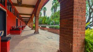 Un pasillo cubierto con paredes rojas y columnas de ladrillo, que conduce a un jardín con plantas y árboles.
