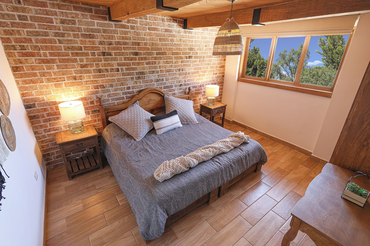 Un dormitorio con pared de ladrillo, suelo de madera, una cama con manta, dos mesitas de noche con lámparas y una ventana con vistas a árboles.