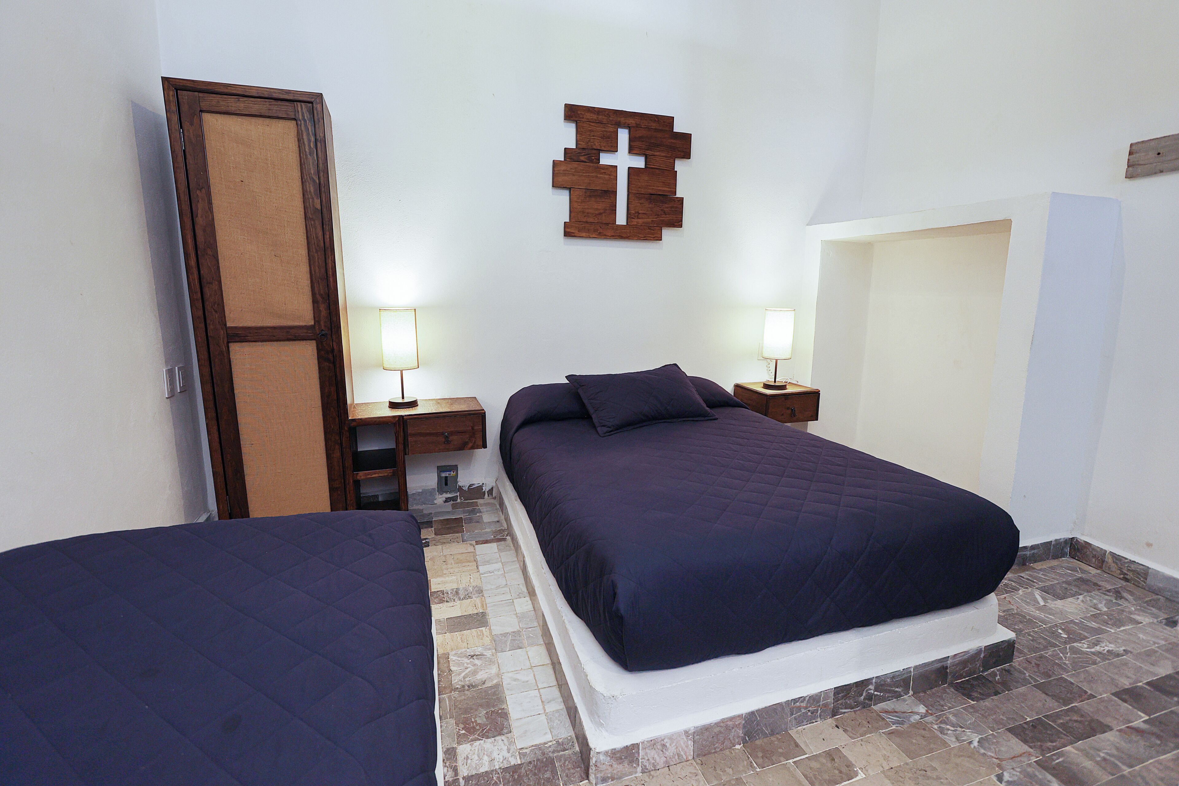 Un dormitorio con dos camas, un armario de madera, un adorno en forma de cruz en la pared y dos lámparas de noche.