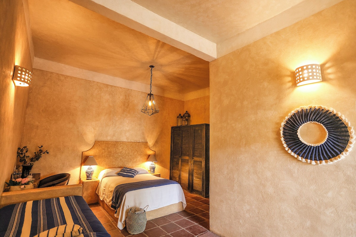 Un dormitorio con cama, lámparas de noche, un candelabro colgante, un armario y un espejo decorativo en la pared.