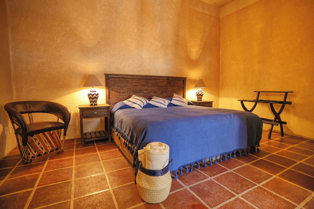 Un dormitorio con cabecera de madera, una cama con cubrecama azul, dos mesitas de noche con lámparas, una silla y una pequeña mesa.
