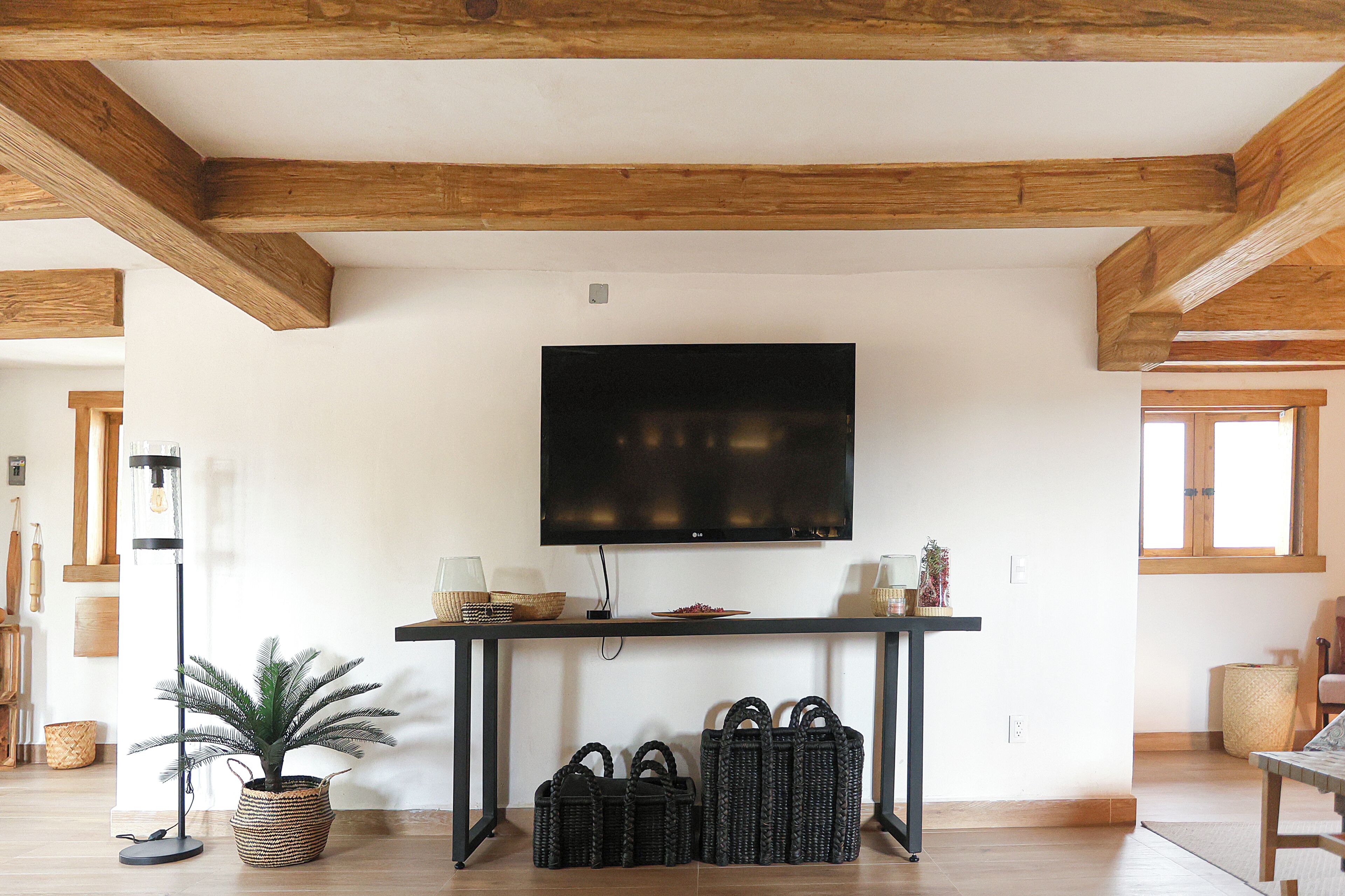 Une pièce avec un plafond en bois, un téléviseur à écran plat fixé au mur, une table avec des paniers et une plante en pot, et un lampadaire.