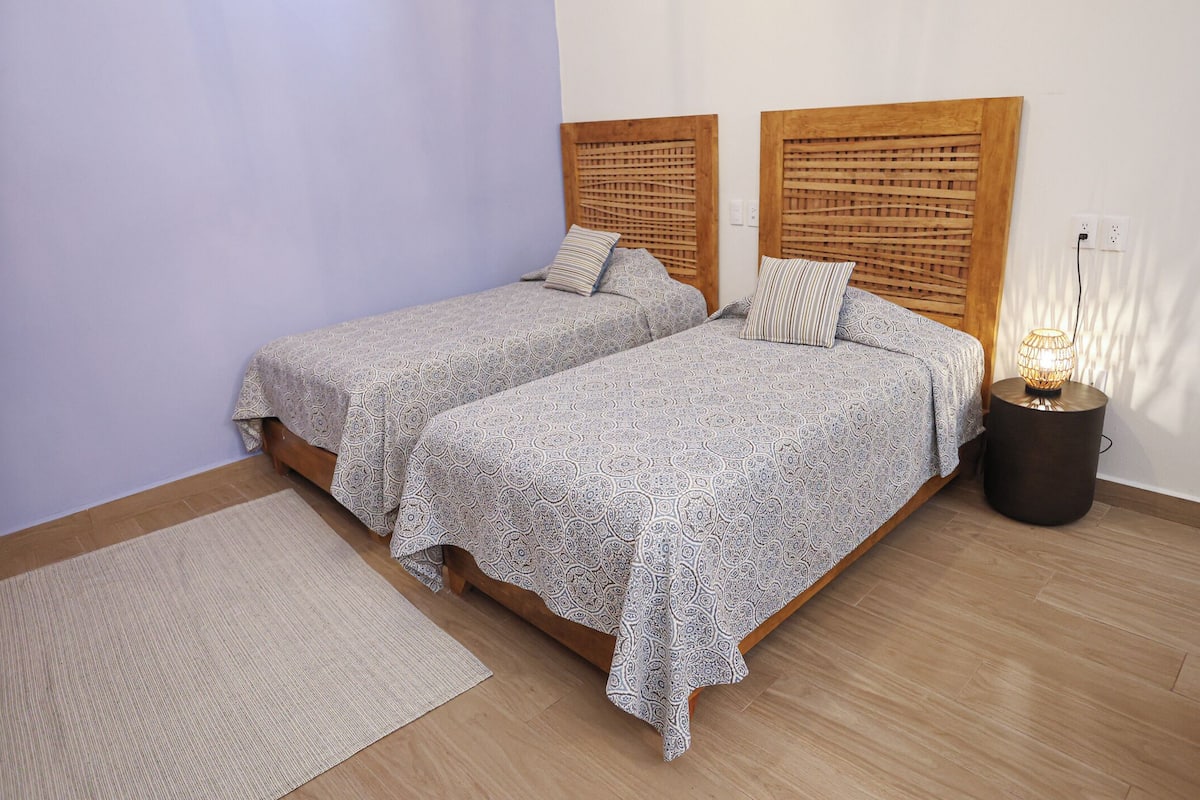 Dos camas con ropa de cama estampada, una mesita redonda con una lámpara y una alfombra sobre piso de madera.