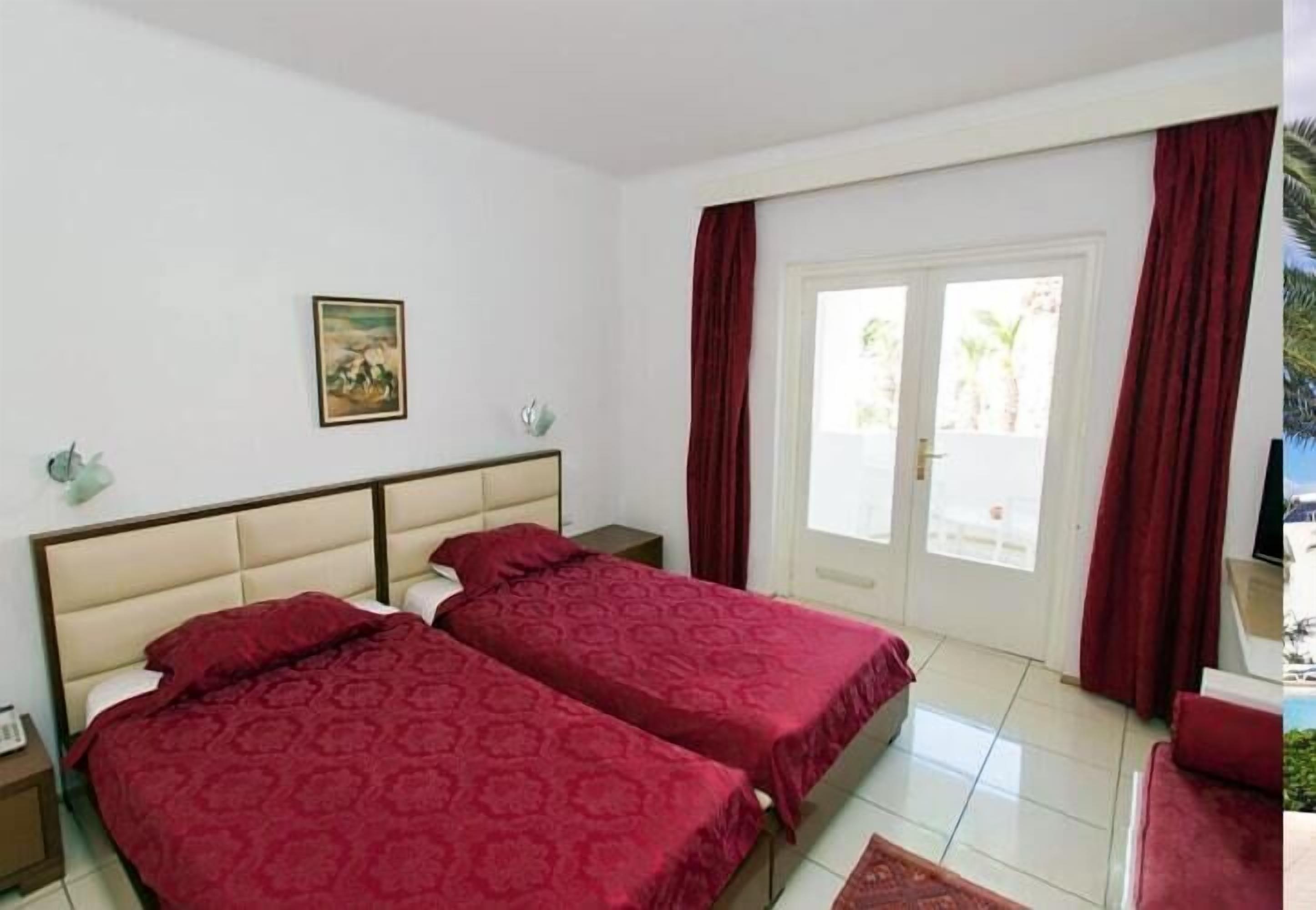 Habitación de hotel con dos camas, puerta al exterior, una mesita y un cuadro en la pared.