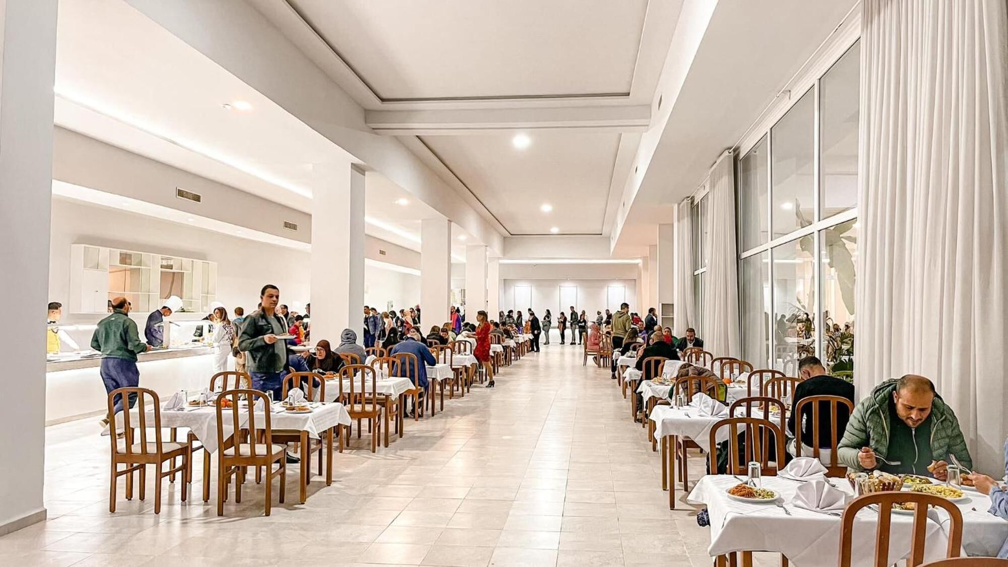 A spacious, modern dining area with long tables and chairs, large windows, and a clean, minimalist design.