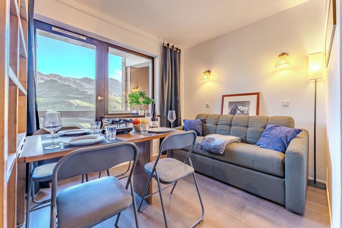 A dining area with a table set for two, a sofa, and a view of mountains through a window.
