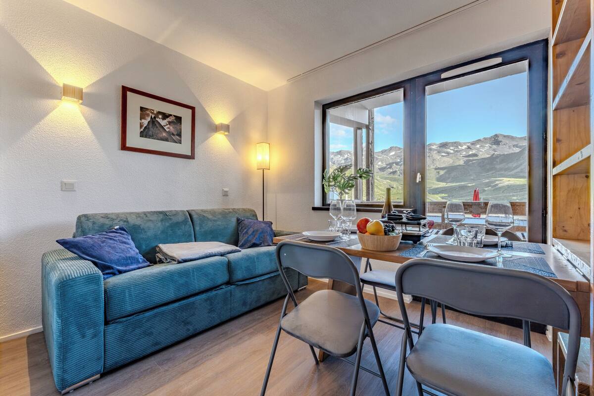 A cozy room with a sofa, dining table, chairs, and a large window offering a mountain view.