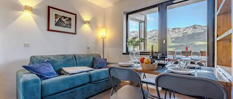 A cozy room with a sofa, dining table, chairs, and a large window offering a mountain view.