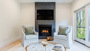 Een moderne woonkamer met twee lichtgrijze fauteuils, een ronde salontafel met een decoratieve schaal, een open haard en een groot raam met uitzicht op groen.