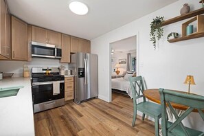 A compact kitchen with modern appliances, a dining area with a wooden table and chairs, and a visible bedroom with a bed and window.