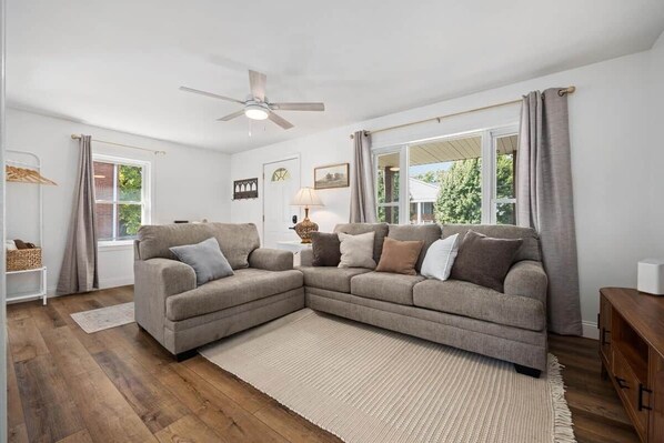 A living room with a sectional sofa, a ceiling fan, a window with curtains, and a wooden floor.