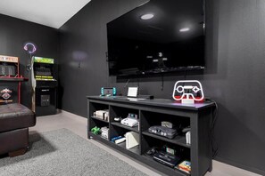 A room with a flat-screen TV mounted on the wall, a gaming console on a shelf, and arcade machines.