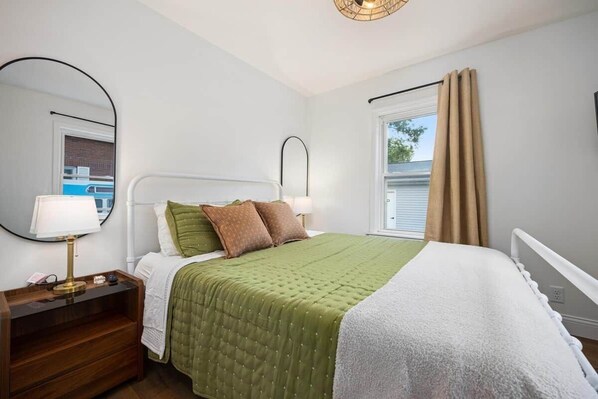 A bedroom with a bed, bedside table, lamp, and a window with curtains. - Play & Stay in the Heights! (Peoria Heights)