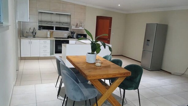 Una cocina moderna con armarios blancos, horno empotrado y ventana con persianas. Hay un comedor con una mesa de madera y dos sillas.