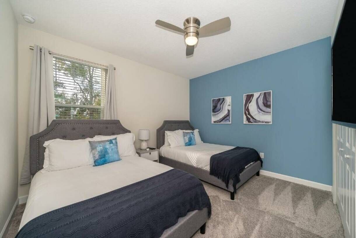 A bedroom with two beds, a ceiling fan, a window with curtains, and two wall art pieces.