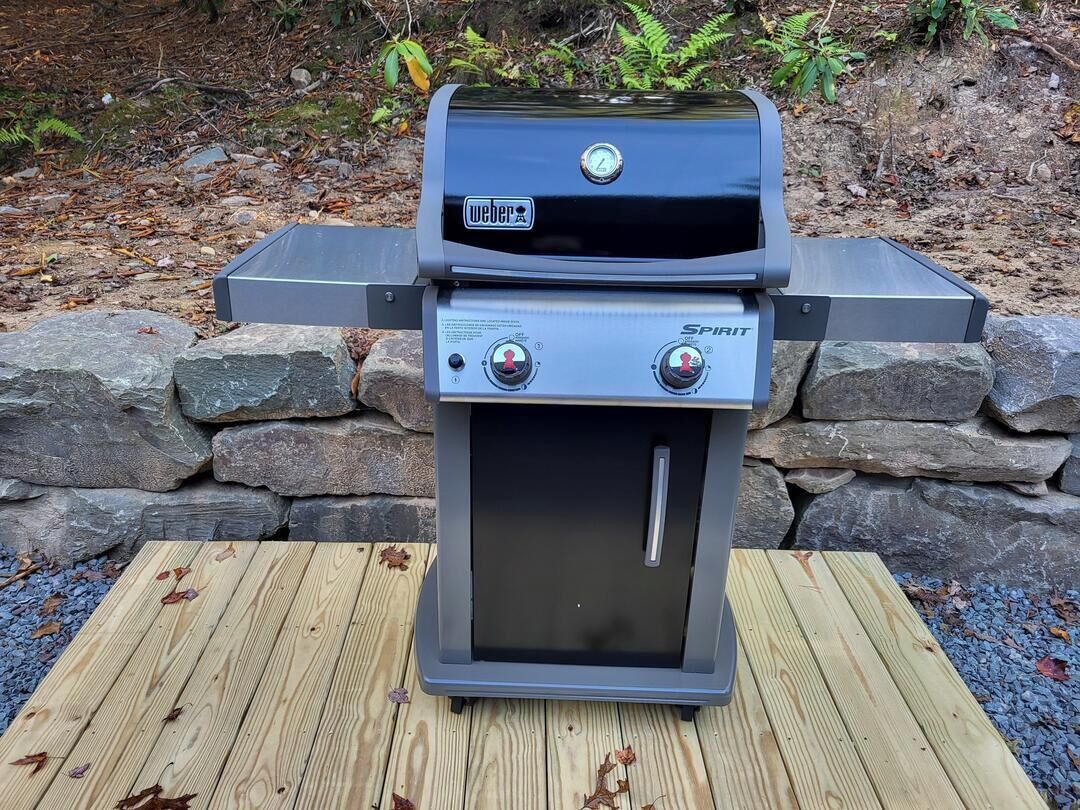 A Weber Spirit portable grill with digital controls, placed on a wooden deck with a stone wall background.