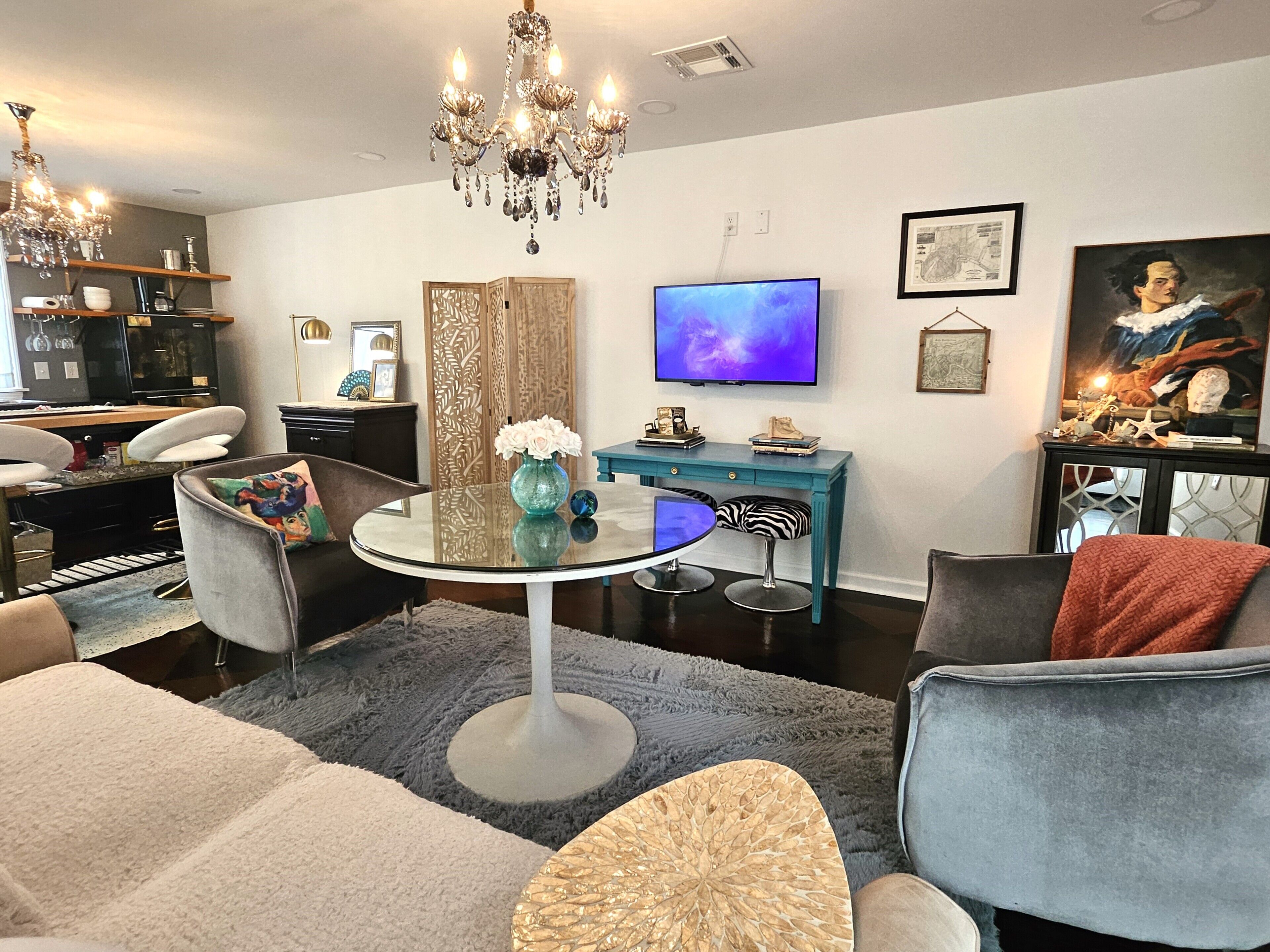 Un salon moderne avec une table ronde en verre, deux fauteuils gris, un canapé, une table d’appoint de couleur turquoise, un téléviseur et un lustre.