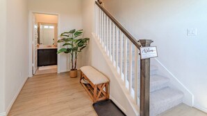 Uma escada de madeira com corrimão branco e um letreiro de boas-vindas, que leva a um quarto com piso de madeira e planta em vaso.