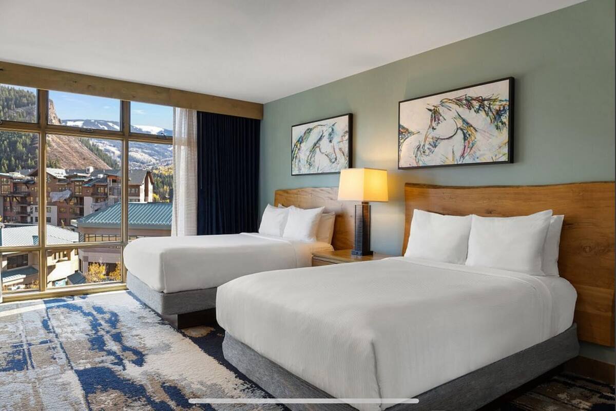 A hotel room with two beds, a large window with a view of mountains, and artwork on the walls.