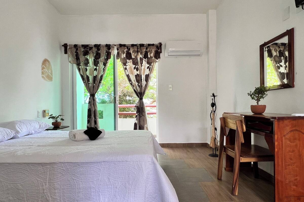 Quarto de hotel com uma cama, uma mesa, uma cadeira, um vaso de planta e vista para o exterior.