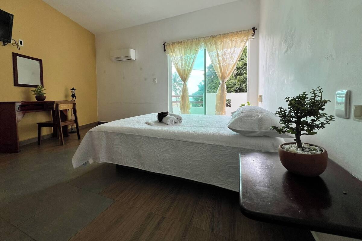 Quarto de hotel com uma cama, escrivaninha, planta em vaso e janela com cortinas.