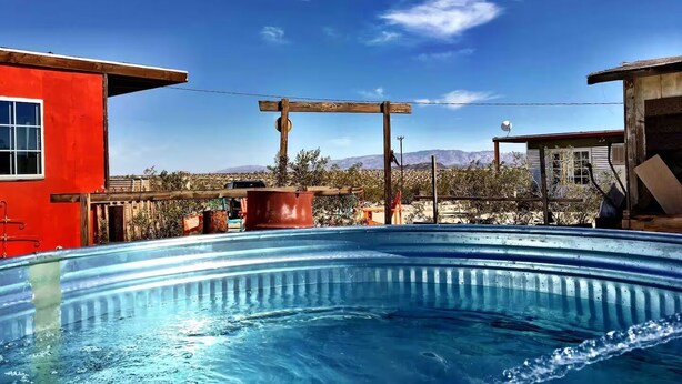 Una piscina con una pérgola de madera y un edificio rojo al fondo.