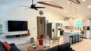 A modern living room with a black sofa, a coffee table, and a TV mounted on the wall. There is a kitchen area with white cabinets and a large island. - New & Cozy 5 BR Lake Anna Retreat - Large Game Room & Lake Access (Bumpass)