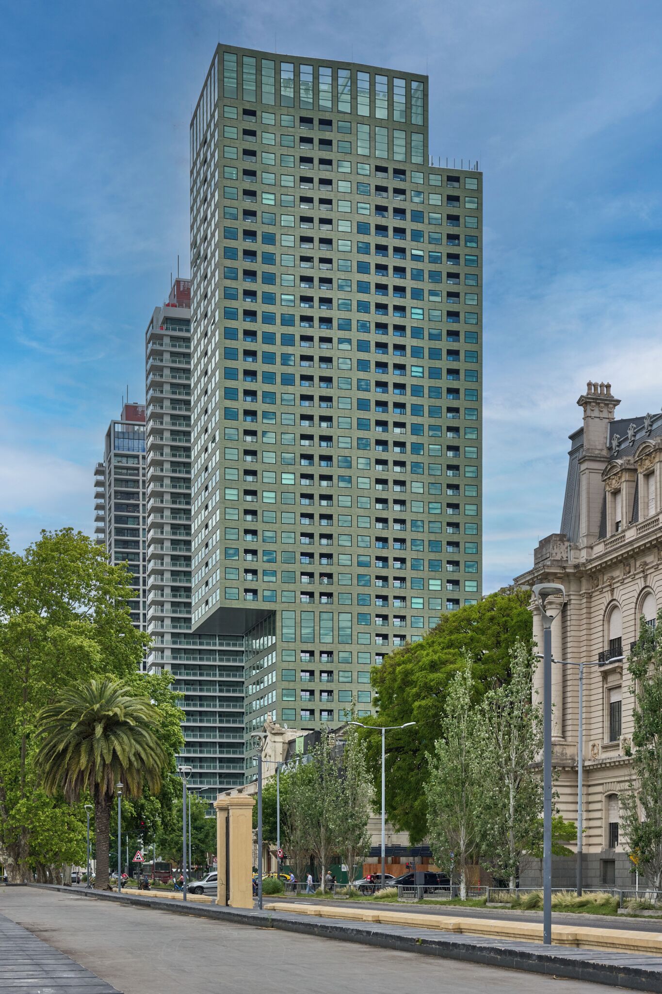 Ein modernes Hochhaus mit einem Raster aus Fenstern, umgeben von Bäumen und einem historischen Gebäude im klassischen Stil.
