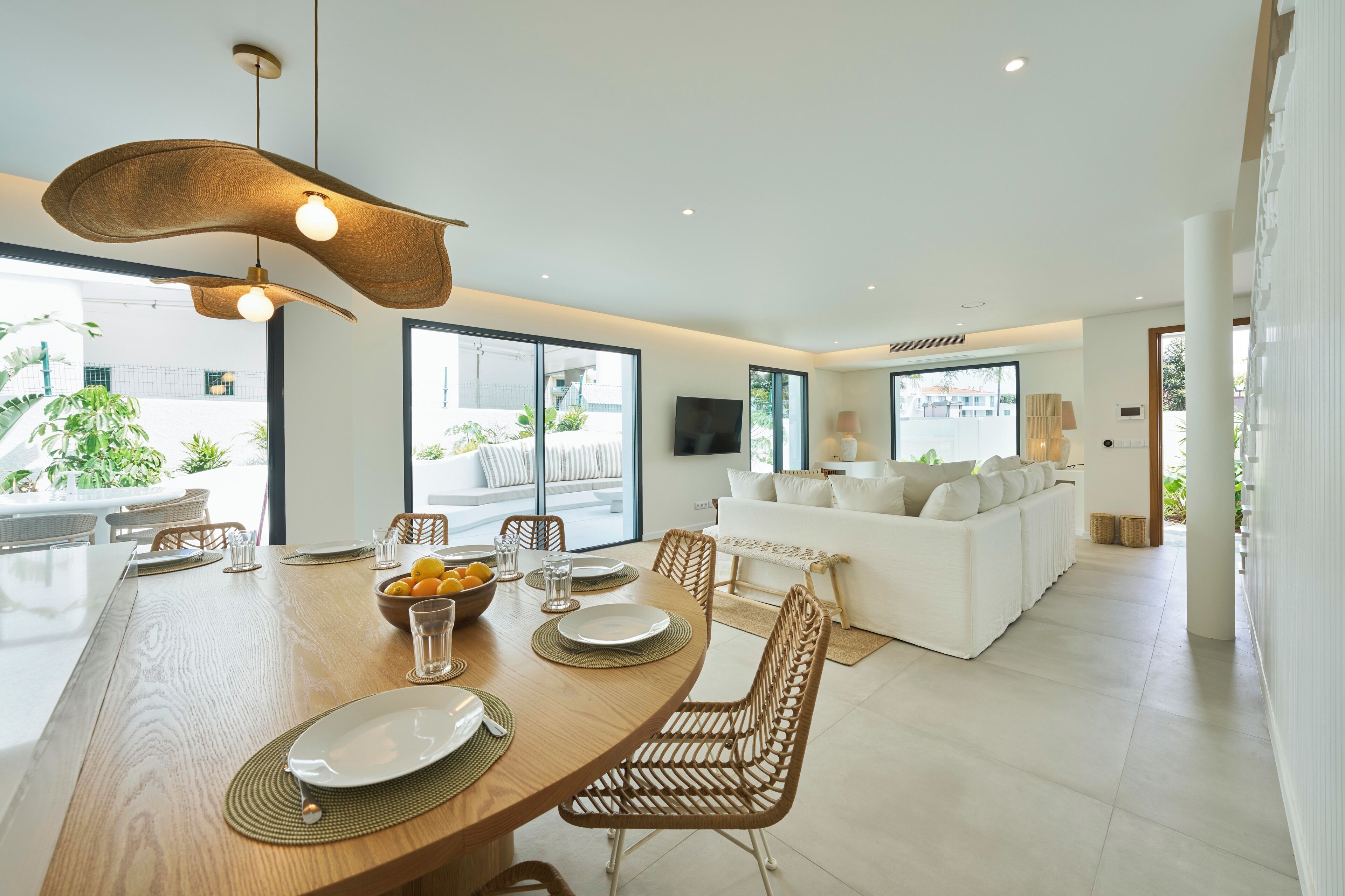 A modern dining area with a wooden table set for four, surrounded by wicker chairs. A white sofa and a flat-screen TV are visible in the background.