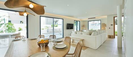 A modern dining area with a wooden table set for four, surrounded by wicker chairs. A white sofa and a flat-screen TV are visible in the background.