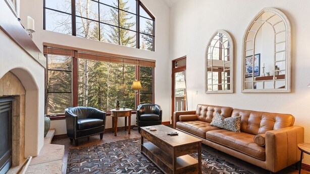 A spacious living room with a fireplace, leather sofas, a coffee table, and large windows offering a view of trees. - Arrowhead at Beaver Creek, Direct Ski Access & A/C (Edwards)