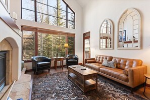 A spacious living room with a fireplace, leather sofas, a coffee table, and large windows offering a view of trees. - Arrowhead at Beaver Creek, Direct Ski Access & A/C (Edwards)