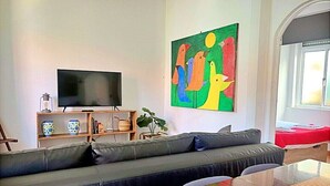 A modern living room with a large flat-screen TV, a colorful abstract painting, a sofa, a wooden coffee table, and a window with a view.