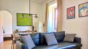 A modern living room with a black leather sofa, a dining table with chairs, a green abstract painting, and a refrigerator.