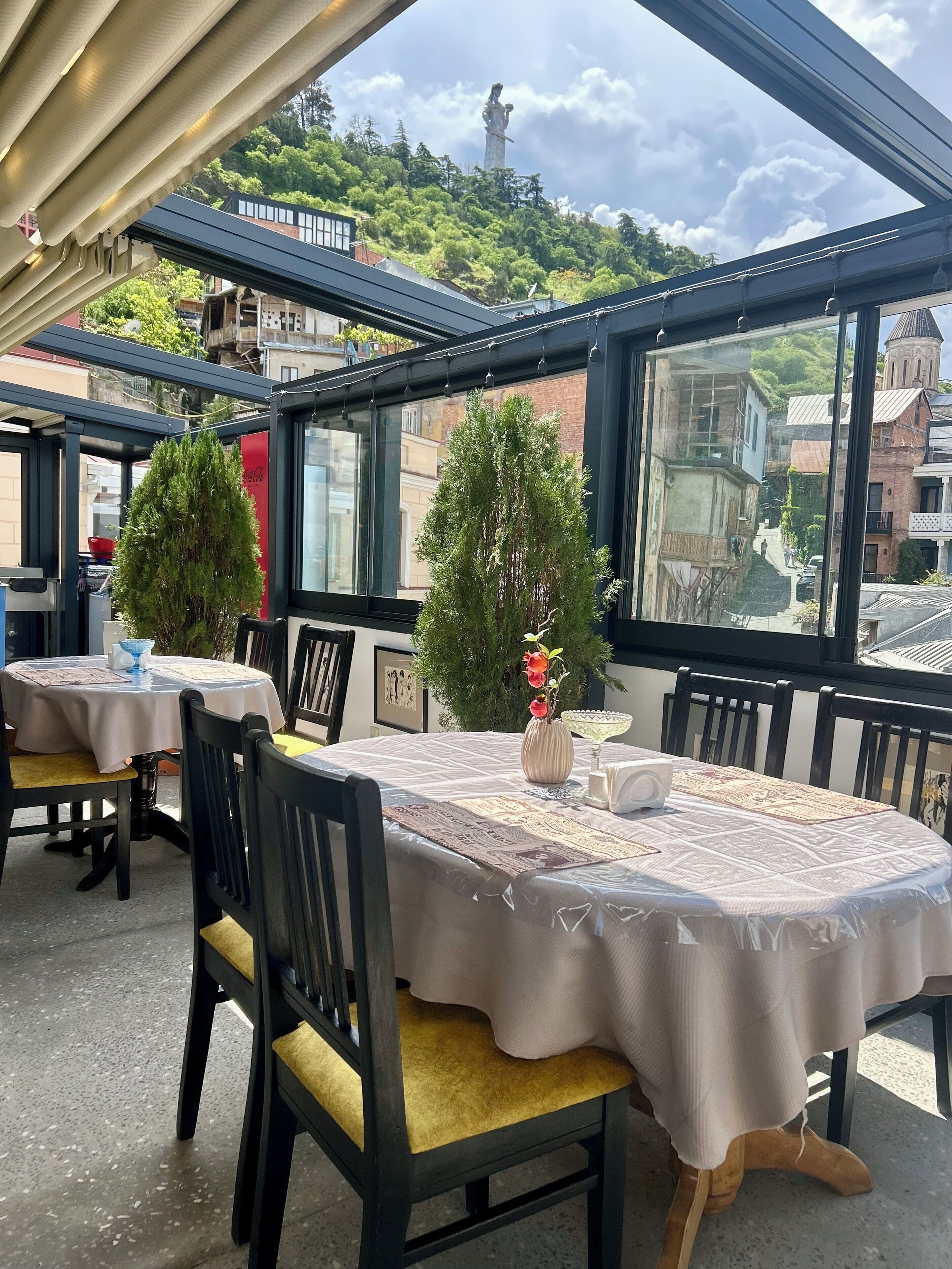 An outdoor dining area with tables covered in white tablecloths, chairs with yellow cushions, and potted plants.