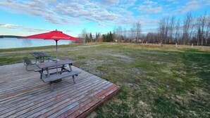 Eine Holzterrasse mit Picknicktisch und einem roten Sonnenschirm, mit Blick auf eine Wiese mit Bäumen und ein Gewässer im Hintergrund.