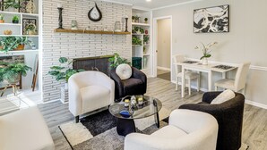 Una sala de estar moderna con chimenea de ladrillo, sofás blancos, una mesa de centro de vidrio y un comedor con mesa y sillas blancas.