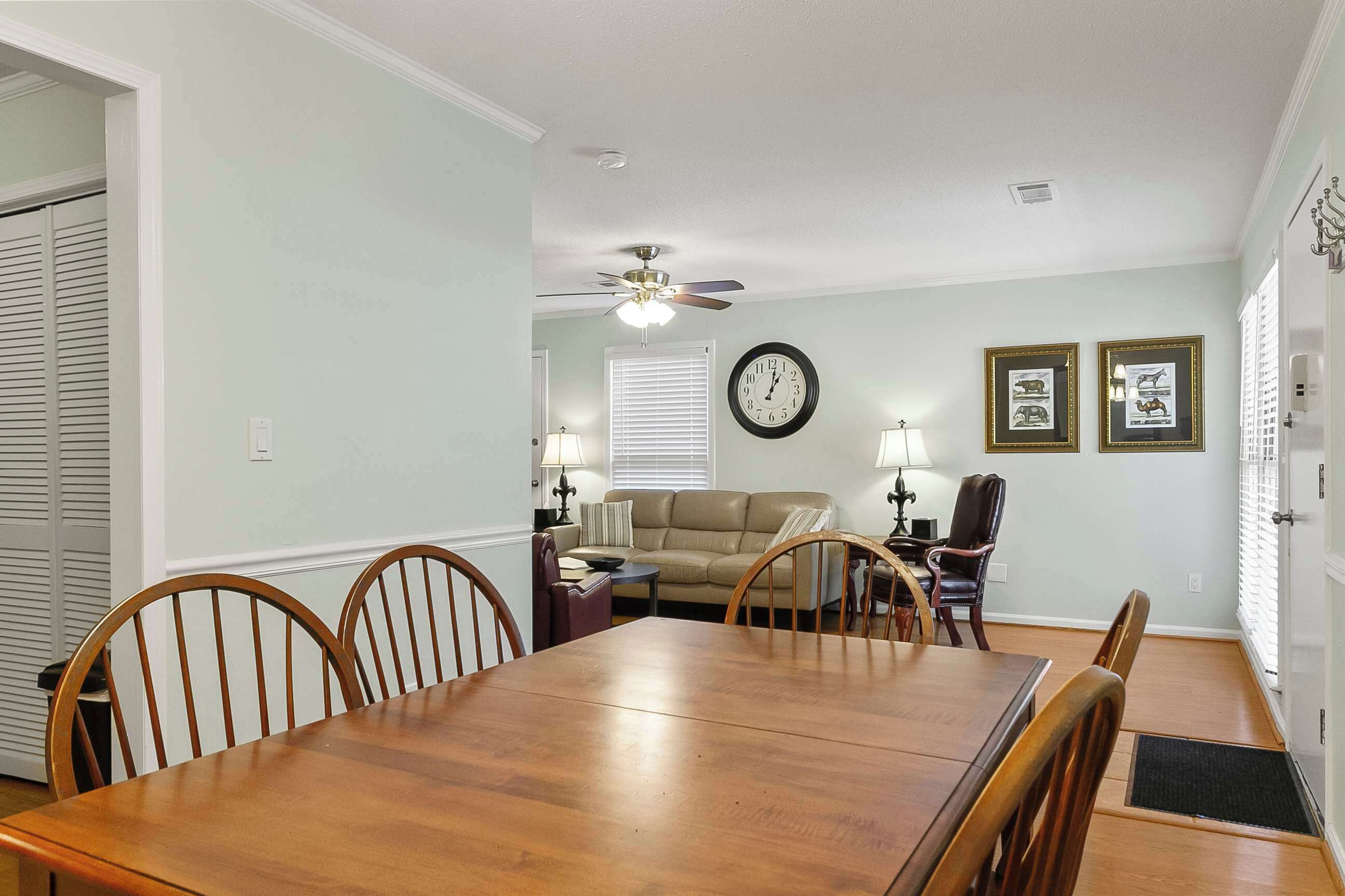 Un coin repas avec une table et des chaises en bois, un ventilateur de plafond, une horloge et des tableaux encadrés accrochés au mur.