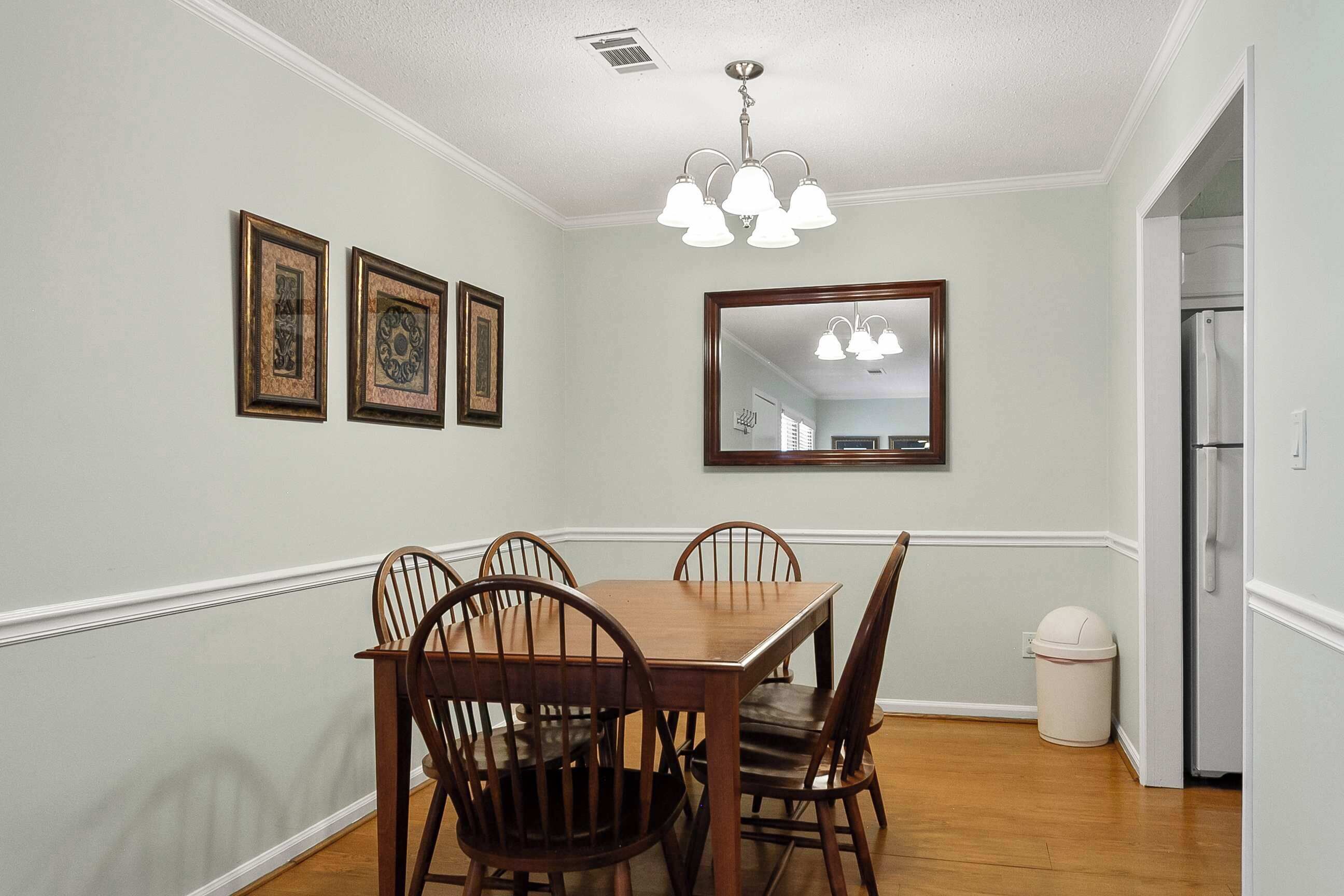A dining room with a wooden table and chairs, a chandelier, framed pictures on the wall, and a trash can.