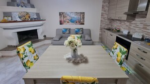 A modern living room with a fireplace, a dining table with floral chairs, a grey sofa, and a kitchen with wooden cabinets and a tiled backsplash. - Giordano Rooms Amalfi Coast  (Tramonti)