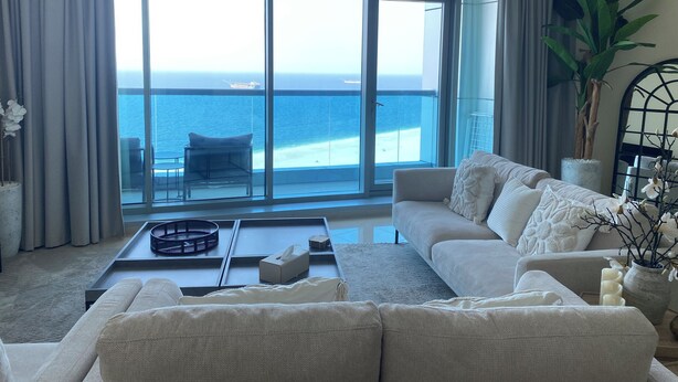 Ein Wohnzimmer mit einem großen Fenster mit Blick auf Strand und Meer. Es gibt ein Sofa, einen Couchtisch und einen Blumentopf.