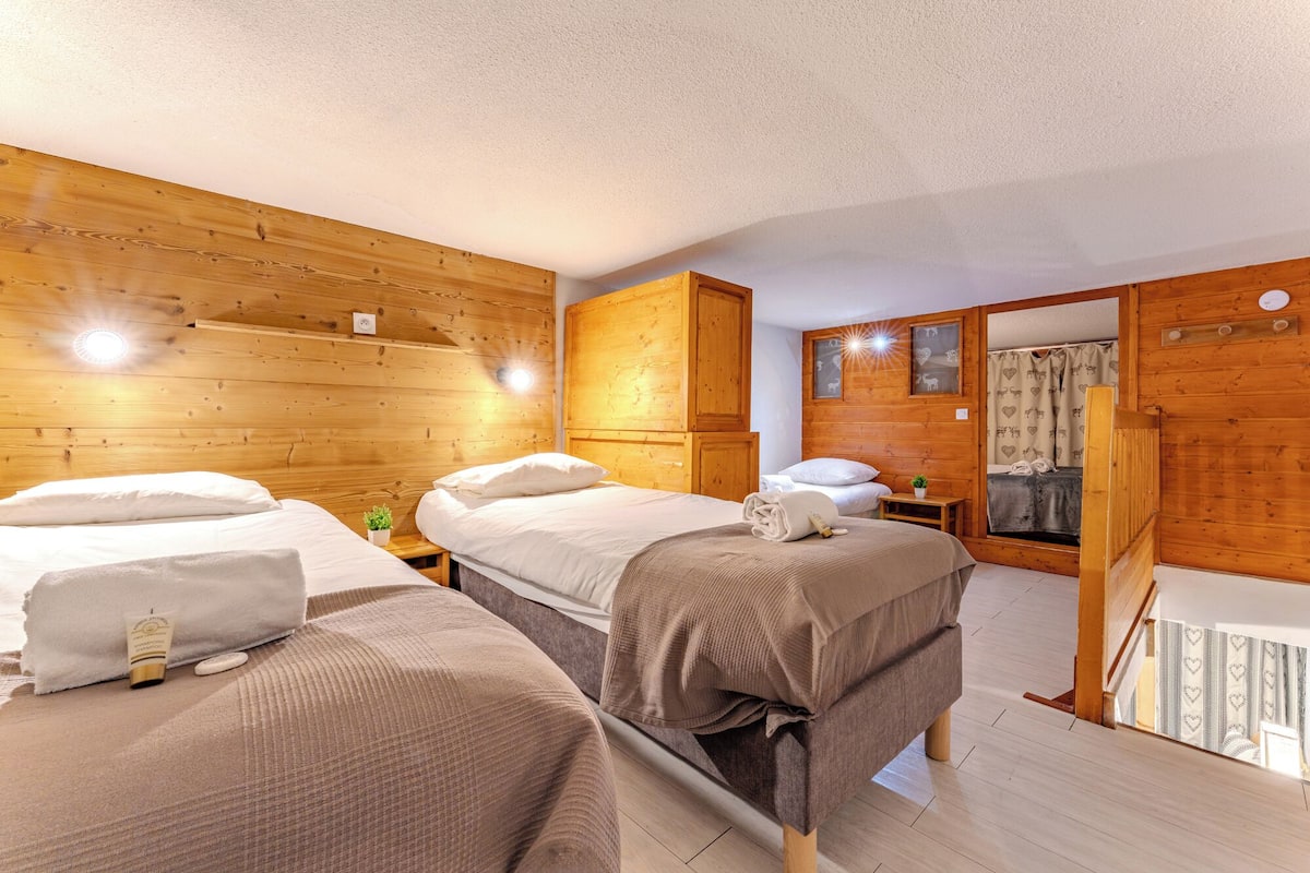 Une pièce chaleureuse avec des murs en bois et un lit recouvert d’une couverture marron. On y trouve une petite table sur laquelle sont posés une plante et un flacon de shampoing.